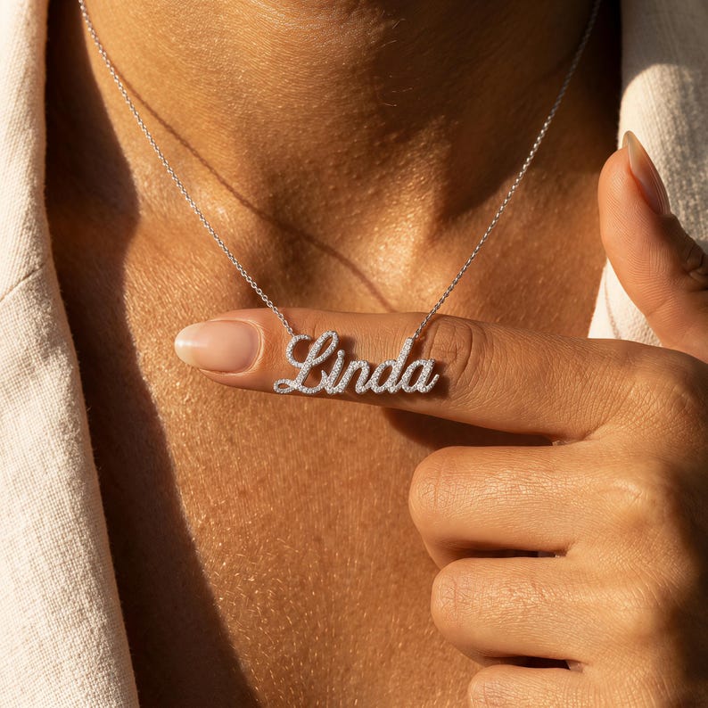 May include: A silver-toned necklace featuring a cursive nameplate pendant that spells out the name Linda. The pendant is encrusted with small, clear, sparkling stones along the entire surface of the letters and hangs from a delicate chain.