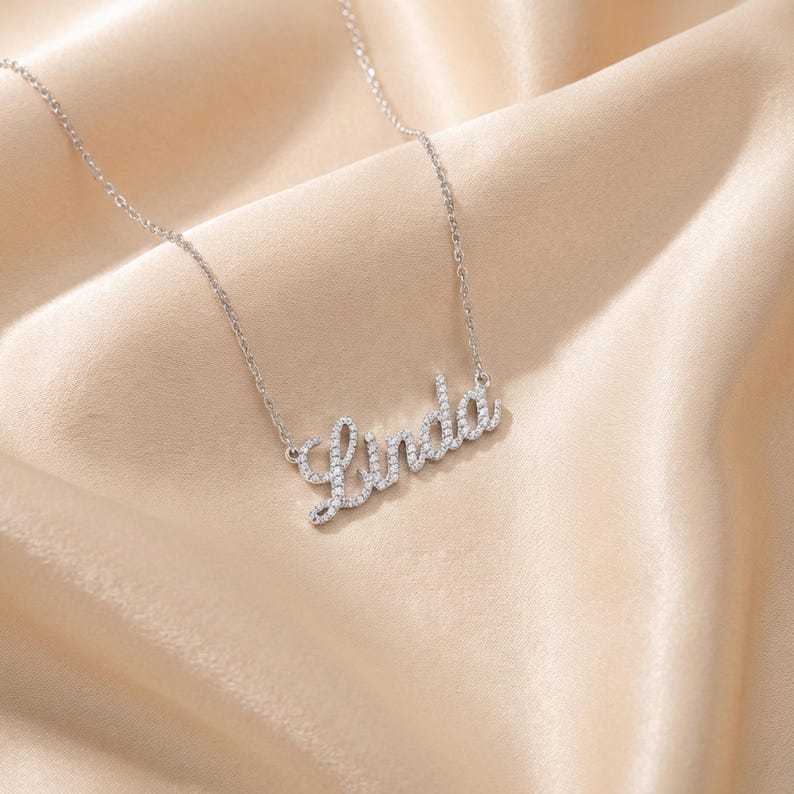 May include: A silver-toned necklace featuring a cursive nameplate pendant that spells the name Linda. The pendant is encrusted with small, clear, sparkling stones along the entire surface of the letters, attached to a delicate link chain.