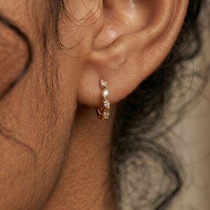 Puede incluir: Un pendiente de aro dorado con varios diamantes pequeños y brillantes. El pendiente se lleva en la oreja, mostrando el delicado diseño y la forma en que los diamantes captan la luz. Una joya clásica y elegante.