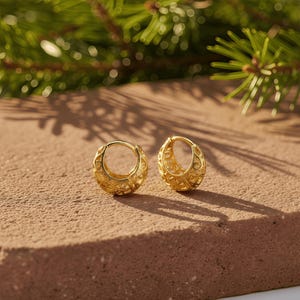 May include: Two gold-colored hoop earrings with intricate filigree patterns. The earrings are round and have a hinged closure. They are displayed on a textured, reddish-brown surface, with evergreen branches in the background.