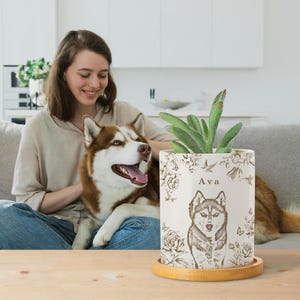 May include: A cream-colored ceramic planter with a detailed illustration of a husky dog, surrounded by floral and bird designs. The planter has the name "Ava" printed on it and sits on a round wooden tray. A green plant is inside.