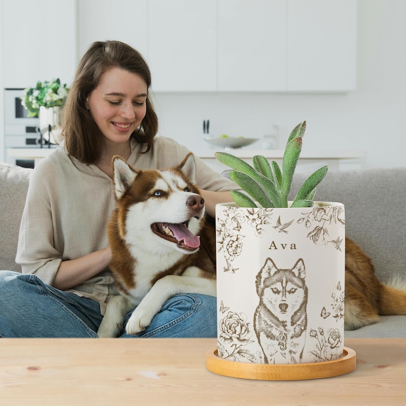 May include: A cream-colored ceramic planter with a detailed illustration of a husky dog, surrounded by floral and bird designs. The planter has the name "Ava" printed on it and sits on a round wooden tray. A green plant is inside.