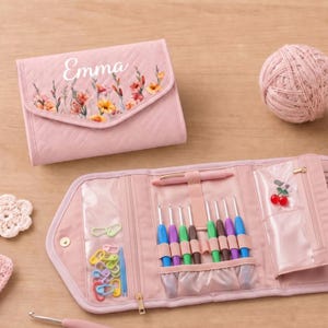 May include: A quilted pink organizer case with the name Emma and a floral print on the flap. The open case reveals clear pockets containing colorful plastic stitch markers, a set of crochet hooks in various colors, and a ball of pink yarn.