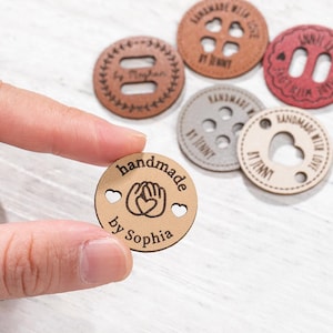 May include: A round, tan leather tag featuring the text "handmade by Sophia" and a graphic of two hands forming a heart shape. The tag has two small heart-shaped cutouts on the sides. Other similar circular leather tags in brown, red, and grey are visible.
