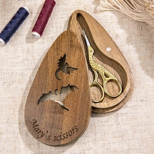 May include: A teardrop-shaped wooden case with a cutout of a dragon and the engraved text "Mary's scissors." Inside the case rests a pair of gold-coloured metal stork-shaped embroidery scissors with intricate detailing on the handles and blades.