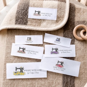 Puede incluir: Una colección de etiquetas rectangulares de tela blanca con una ilustración de máquina de coser vintage negra, detalles florales y el texto impreso Made With Love seguido de varios nombres, dispuestas sobre una manta de lana a cuadros beige y marrón.