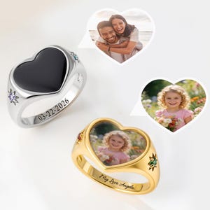 May include: A silver-toned heart-shaped ring with a black inlay and the date 03-22-2026 engraved on the band, alongside a gold-toned heart-shaped ring featuring a photo of a child and the engraved text My Love Angela on the band.