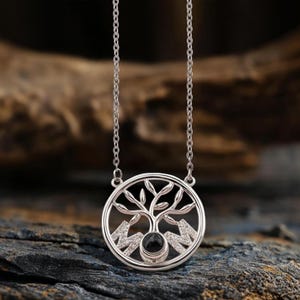 May include: Silver necklace with a circular pendant. The pendant features a tree design with leaves and branches, mountains, and the letters "M" and "M". A black stone is at the centre of the tree design. The necklace hangs on a silver chain.