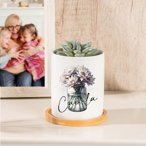 Puede incluir: Maceta de cerámica blanca con base de madera. La maceta presenta un diseño floral de nenúfares blancos en un frasco de vidrio. El texto "Camila" está escrito en cursiva en la maceta.