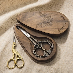 May include: A pair of ornate metal scissors with floral handles resting inside a teardrop-shaped wooden case. The case lid features an engraved butterfly and the text "Mary's scissors". A second pair of gold-coloured bird-shaped scissors sits nearby.