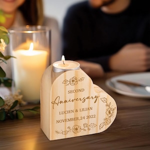 May include: A wooden heart-shaped candle holder with a lit tea light. The holder is engraved with "SECOND Anniversary", names, and a date. Floral designs are etched around the text. A large candle in a glass jar is in the background.