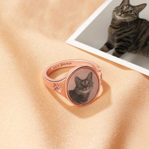 May include: Rose gold ring with a cat portrait and the inscription "2021-forever." The ring features a small star and a purple gemstone. A photograph of the cat is in the background.