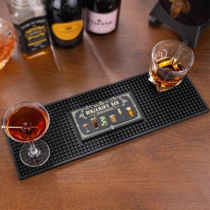 May include: A rectangular black rubber bar mat with a textured nub surface and a central graphic featuring the text "WELCOME TO BENJAMIN'S BAR HOUSE FAVORITES" alongside illustrations of various cocktails, a martini glass, and a beer mug.