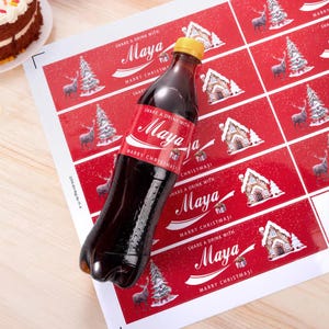 Può includere: Una bottiglia di bibita marrone scuro con tappo giallo e un'etichetta rossa con la scritta "SHARE A DRINK WITH Maya MARRY CHRISTMAS!", decorata con illustrazioni di una casa di pan di zenzero, un albero di Natale e una renna.