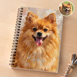 May include: A spiral-bound notebook featuring an oil painting style portrait of a fluffy orange Pomeranian dog with its tongue out. The name Rocky is written in white cursive script across the lower portion of the cover. A small circular inset shows the original dog photo.