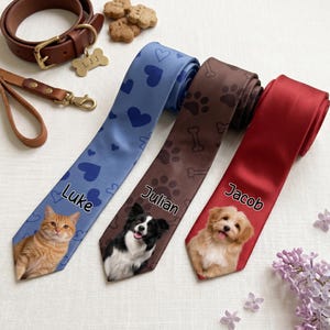 Puede incluir: Tres corbatas con retratos de mascotas y nombres personalizados. La corbata azul tiene corazones y un gato llamado Luke, la marrón huellas y huesos con un perro llamado Julian, y la roja muestra un perro llamado Jacob. Incluye un collar de cuero.