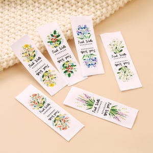 May include: Six rectangular white fabric labels featuring colorful floral illustrations and the black printed text Trish Wells Handmade. Each label displays a unique botanical design, including holly, wildflowers, and lavender, arranged in a vertical layout.