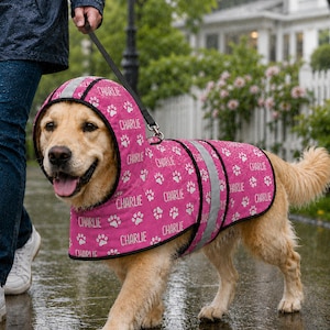 Könnte beinhalten: Ein rosafarbener Hunde-Regenmantel mit Kapuze, der mit einem sich wiederholenden Muster aus weißen Pfotenabdrücken und dem in Weiß gedruckten Namen Charlie versehen ist. Das Kleidungsstück hat reflektierende silberne Streifen und eine Metallöse.