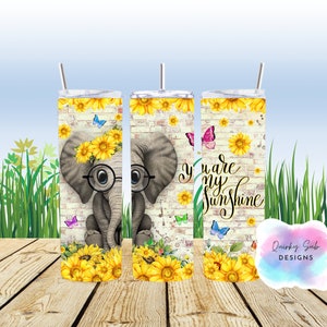 May include: A white tumbler with a design of a cartoon elephant wearing glasses and a sunflower crown. The background is a brick wall with sunflowers and butterflies. The text "You are my sunshine" is on the tumbler.