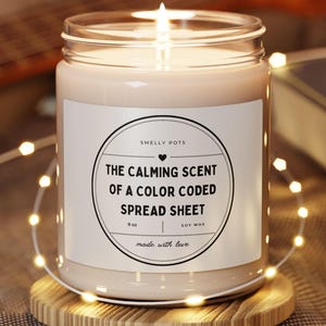 Puede incluir: Una vela de cera de soja encendida en un tarro de cristal con una etiqueta blanca que dice "The Calming Scent of a Color Coded Spreadsheet". La vela está sobre un pequeño posavasos de madera, rodeada de luces de cadena.