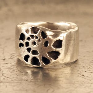 May include: A silver ring featuring a textured, spiral design. The ring is wide and has a rounded, slightly irregular shape.