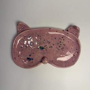 Può includere: Un piatto rosa in ceramica a forma di maschera per dormire con vernice maculata. Il piatto ha due sporgenze a forma di orecchio e un piccolo nasello arrotondato. La vernice maculata include punti neri, blu e verdi.