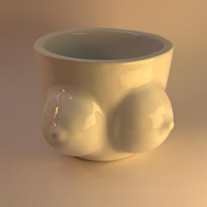 May include: A white ceramic mug featuring a sculptural design on the exterior with two rounded, breast-shaped protrusions. The mug has a smooth, glossy finish and a wide, open top, showcasing a clean, minimalist aesthetic in a solid white color.