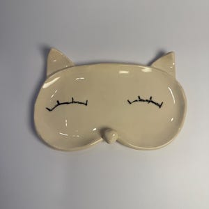 Può includere: Un piatto in ceramica color crema a forma di gatto con occhi chiusi ricamati in nero e un piccolo naso. Il piatto ha due sporgenze a forma di orecchio nella parte superiore. Il design è semplice ed elegante, adatto a contenere piccoli oggetti.