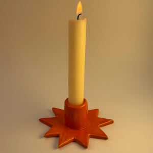 May include: A bright orange ceramic candle holder shaped like a multi-pointed star with a central cylindrical socket. A tall, off-white taper candle is inserted into the holder, featuring a small, flickering flame at the top of the wick.