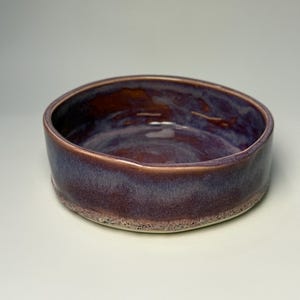 Könnte beinhalten: Eine runde Keramikschale mit flachem Profil und einer glatten, glänzenden Innenglasur in tiefen Violett- und Brauntönen. Die Außenseite weist eine texturierte, gesprenkelte Basis auf, die zum Rand hin in ein glattes, dunkles Violett übergeht.