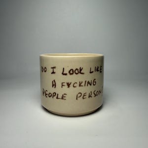 May include: A cylindrical ceramic mug with a smooth, off-white finish. The exterior features dark brown, hand-lettered text that reads: DO I LOOK LIKE A FYCKING PEOPLE PERSON?