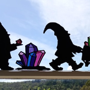 Könnte beinhalten: Silhouetten-Ausschnitte von Zwergen, die bunte Kristalle abbauen. Ein Zwerg hält einen Kristall, ein anderer zieht einen Wagen voller Edelsteine, und ein dritter trägt eine Spitzhacke. Die Kristalle sind in verschiedenen Farben, darunter Lila, Rosa und Grün.