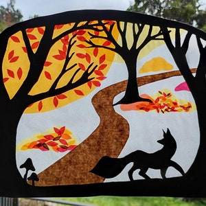 May include: A paper craft depicting a woodland scene with a fox walking along a path. The trees are black silhouettes with yellow, orange, and red leaves. The path is brown and the background is white with a light blue sky.
