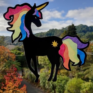 May include: A black unicorn silhouette with a rainbow mane and tail, and a yellow star on its flank. The unicorn is positioned in front of a natural outdoor background with trees and a cloudy sky.