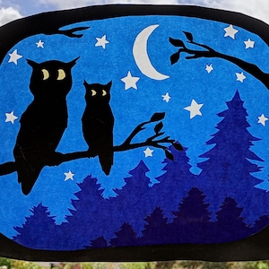 Op de afbeelding: Een kunstwerk met nachtthema met twee uilensilhouetten op een tak tegen een blauwe achtergrond. De scène bevat een halve maan, sterren en een bos-silhouet. Het kunstwerk is omlijst met een zwarte rand.