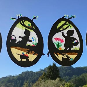 May include: Four oval-shaped window clings featuring black silhouettes of children and rabbits in outdoor scenes. Each egg-shaped design includes colourful paper cutouts of flowers, butterflies, and baskets, creating a layered, translucent effect.