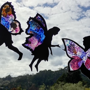 May include: Black silhouette fairy cutouts with stained-glass style wings in shades of blue, purple, and pink. The fairies appear to be in flight against a cloudy sky and a green landscape. The wings are the focal point of the design.