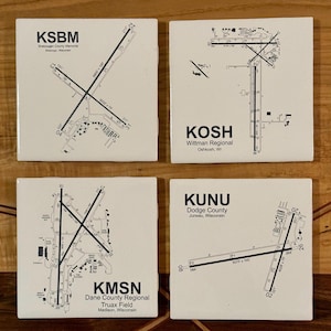 May include: Four white ceramic coasters with black airport runway diagrams and airport codes. The coasters feature the airport codes KSBM, KOSH, KUNU, and KMSN, along with the airport names and locations.