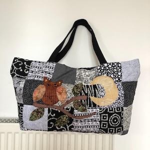 May include: Black and white patchwork tote bag with an owl applique and a crescent moon design. The bag has black straps.