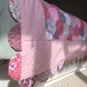 May include: A pink and white patchwork quilt with a scalloped edge. The quilt is made of various floral fabrics and is displayed on a bed.