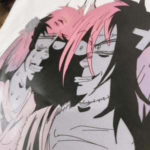 May include: Close-up of a white t-shirt featuring a manga-style illustration. The artwork depicts two characters with pink and black hair, with one character smiling and the other appearing distressed. The design is in shades of grey, pink, and black.