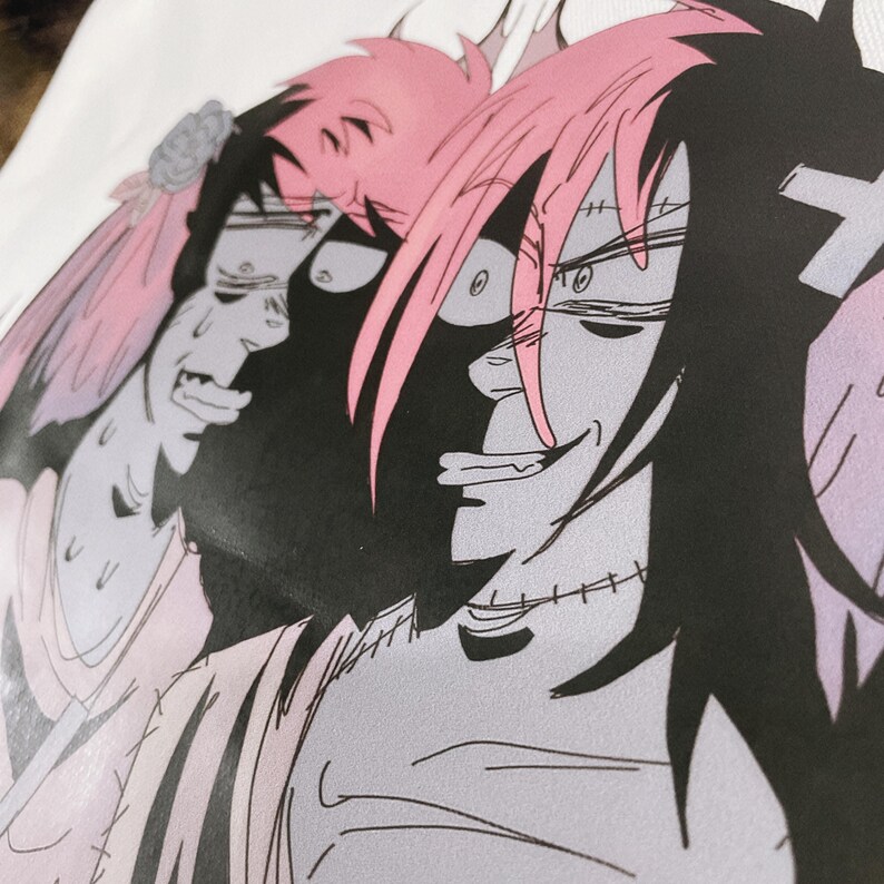 May include: Close-up of a white t-shirt featuring a manga-style illustration. The artwork depicts two characters with pink and black hair, with one character smiling and the other appearing distressed. The design is in shades of grey, pink, and black.