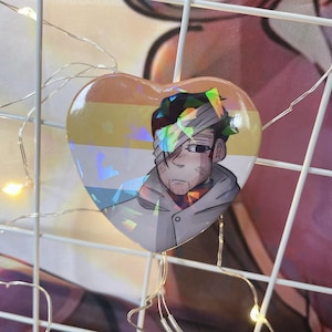 May include: A heart-shaped button with a holographic finish features a cartoon illustration of a person with a bandage over one eye. The person is wearing a gray jacket and has a yellow, white, and blue striped background.