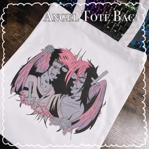 May include: White tote bag featuring a graphic illustration of two figures with angel wings, crosses, and roses. The figures have pink and black hair and are in a distressed style. The text "ANGEL TOTE BAG!" is at the top.