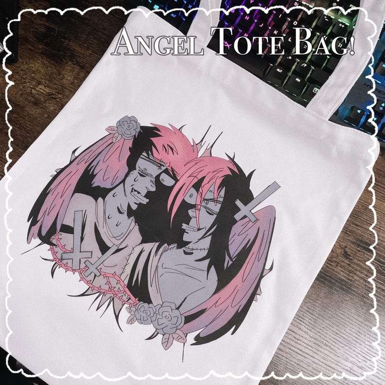 May include: White tote bag featuring a graphic illustration of two figures with angel wings, crosses, and roses. The figures have pink and black hair and are in a distressed style. The text "ANGEL TOTE BAG!" is at the top.