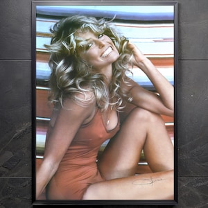 May include: Framed print featuring a smiling person with long, wavy blonde hair. The person is wearing a rust-colored swimsuit and is seated. The background shows a striped pattern in shades of blue, orange, and white. A signature is visible.