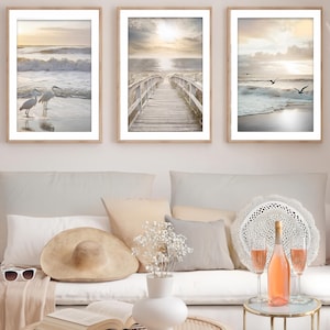 Op de afbeelding: Drie ingelijste strandscènes. De prenten tonen een serene kust met zilverreigers, een houten loopbrug en zeemeeuwen. De kunstwerken worden getoond boven een witte bank met kussens, een zonnehoed, zonnebril en een tafel met rosé en glazen.