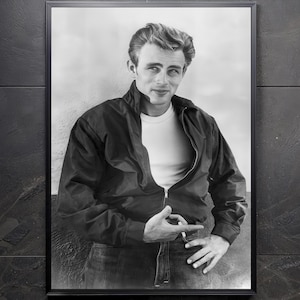 May include: Black and white framed print of a man in a black jacket and white t-shirt. The man is leaning against a wall, with his hand on his hip and holding a cigarette. The print is in a black frame.
