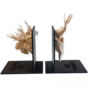 Puede incluir: Un par de sujetalibros negros con esculturas detalladas en marrón claro de una cabeza de criatura y una calavera. La base tiene el texto "THE WITCHER". Diseñados para mantener los libros en posición vertical.