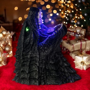 Pode incluir: Uma escultura de cabeça de Godzilla preta com olhos azuis e verdes brilhantes. A escultura está sobre uma superfície vermelha com presentes embrulhados no fundo.